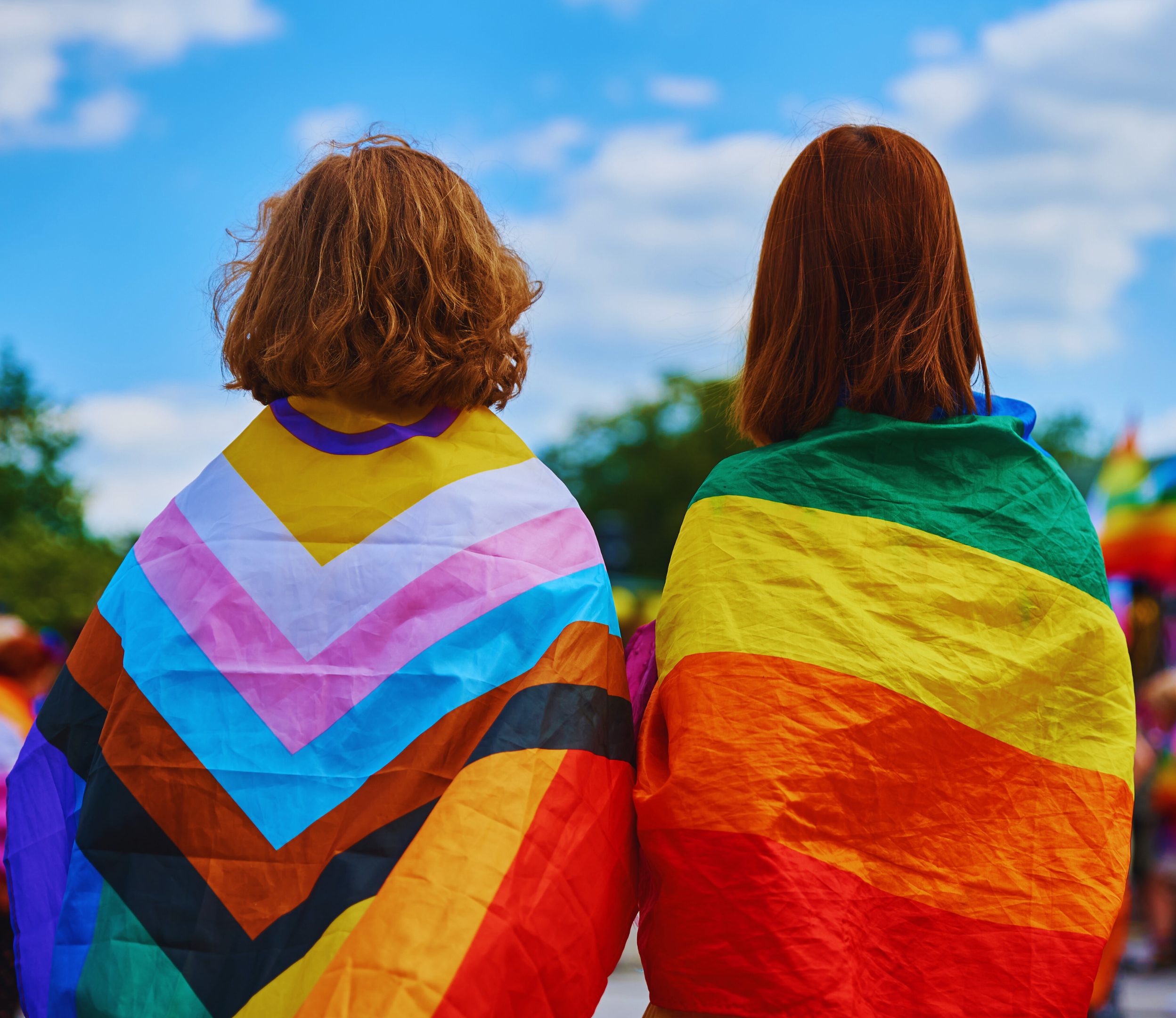 Two people wear pride flags on their backs outside on a sunny day.