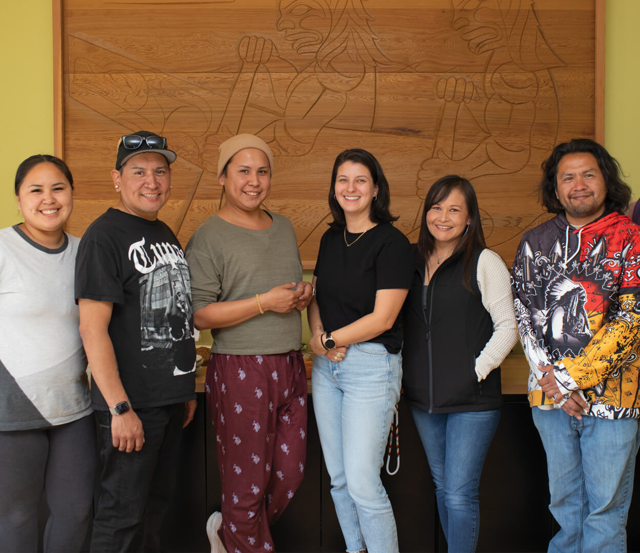 A group of people are smiling at the camera as they stand in front of a large wooden Indigenous artwork.