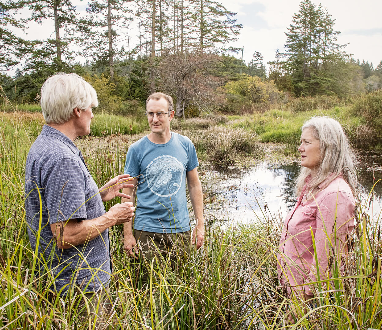 Three people are having a conversation outdoors while standing near marshland.