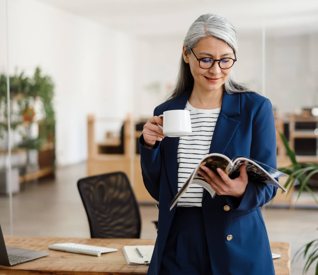 A woman is drinking coffee in her office while reading a printed publication in her hand.