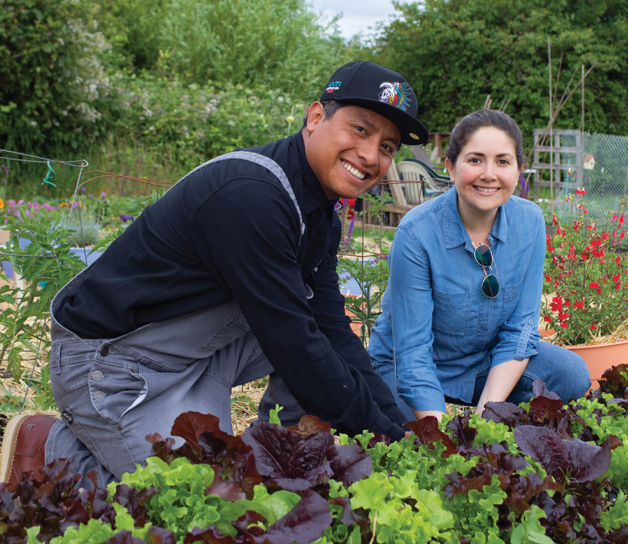 A man and woman smile at the camera while harvesting vegetables in a community garden.